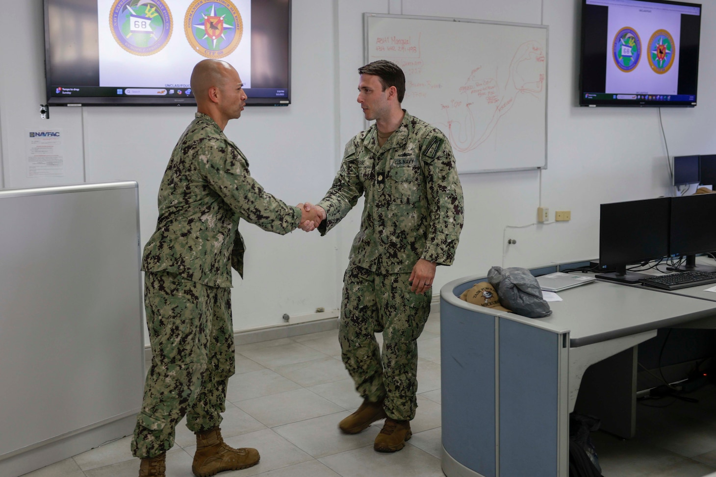 Two Navy personnel shake hands before a brief in a classroom.