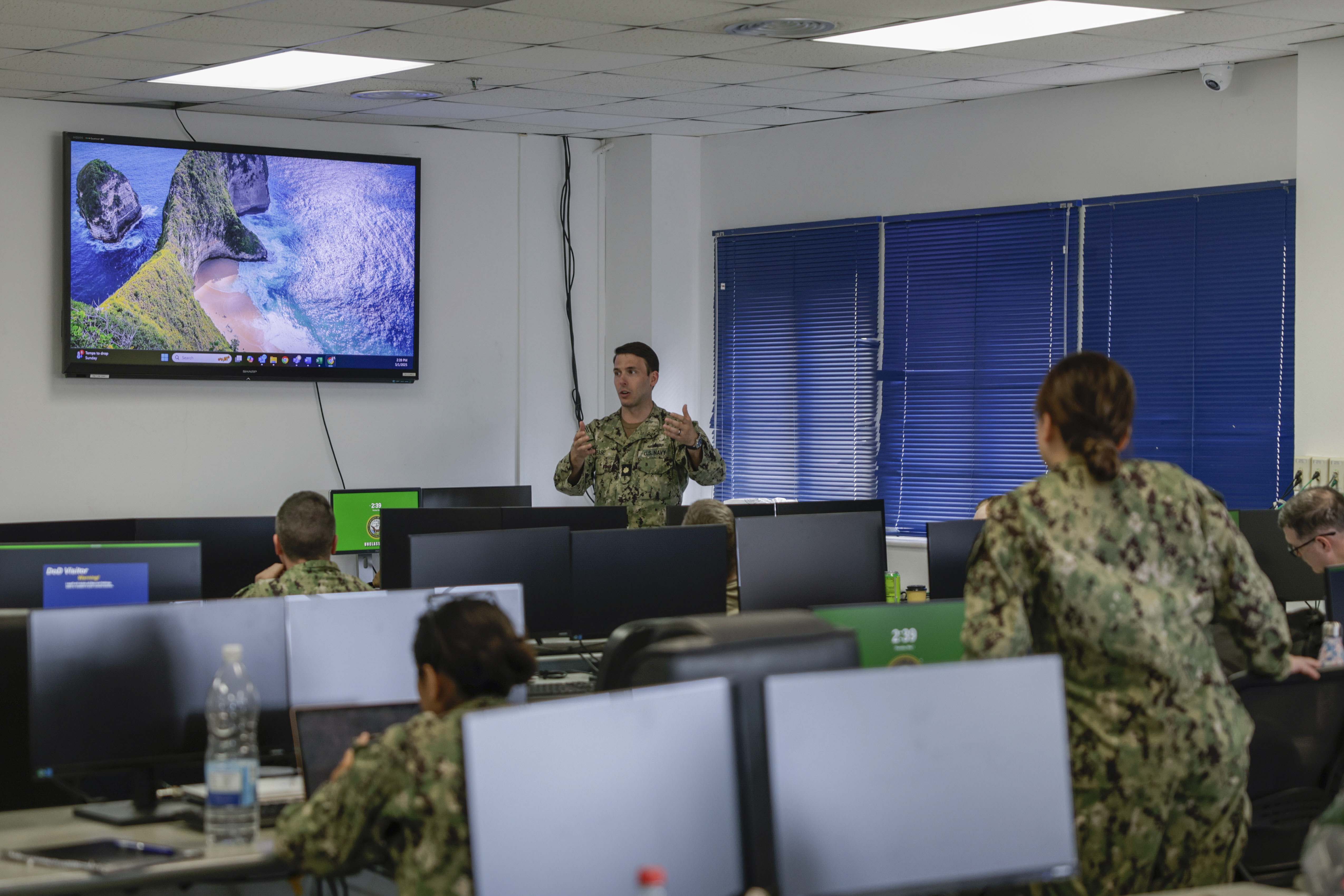 A Navy officer explains something to a class of Navy personnel in a room with computers.