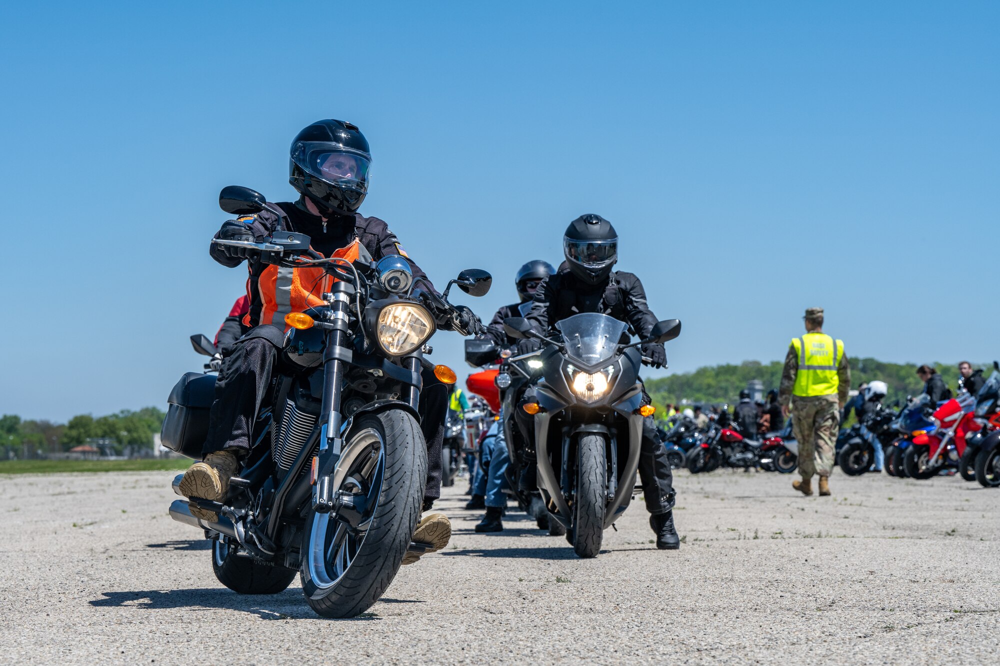 Two motorcycles pointed towards the viewer with headlights on. A row of motorcycles can be seen from the side with a military member wearing a neon yellow safety vest