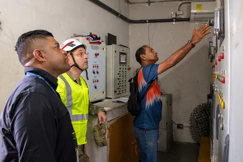 three men, one a Soldier, look at a panel of gauges and switches. The Soldier is wearing a hard hat and protective vest.