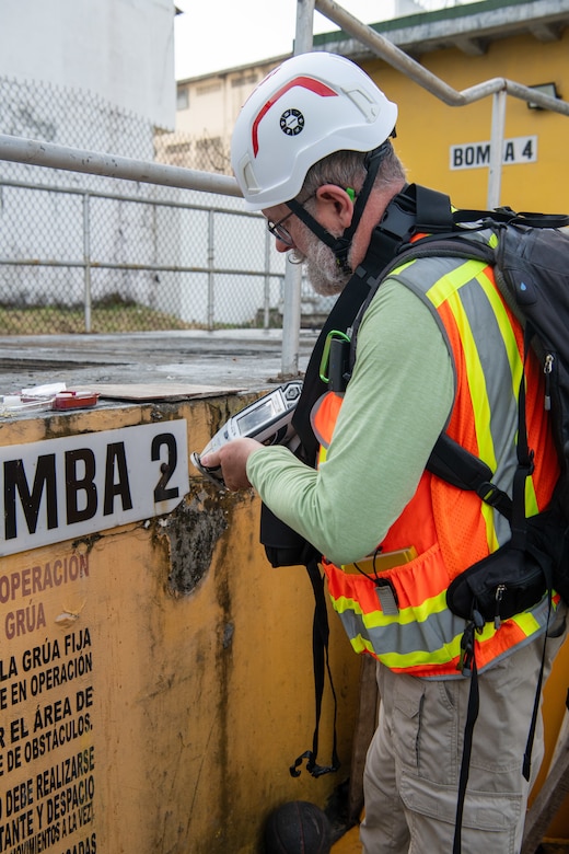 A man wearing a hard hat and reflective vest uses a tool to scan a metal container.