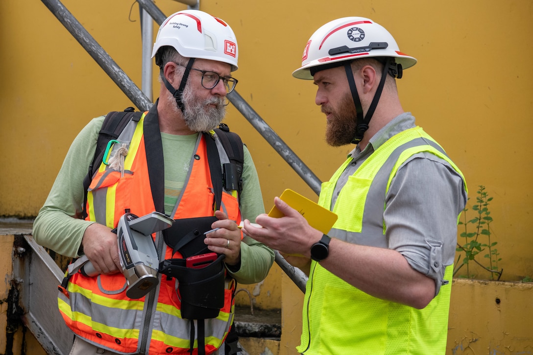 Two men in hard hats and reflective vests are talking to each other.