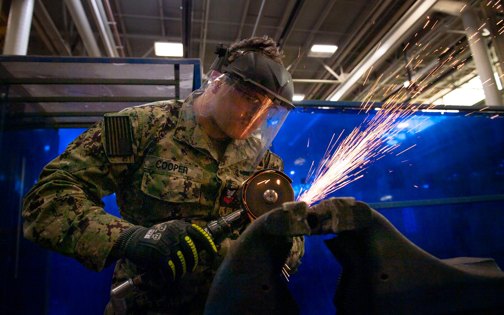 A Navy sailor in a Navy utility uniform welds some metal. There are sparks flying. He has a visor on.