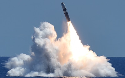 A missile launches from beneath the sea. It is a large tubular vessel with a lot of white capped water surrounding it as it shoots into the clear blue sky.