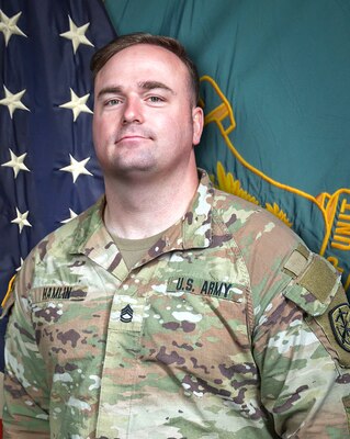 Man in U.S. Army uniform standing in front of two flags.