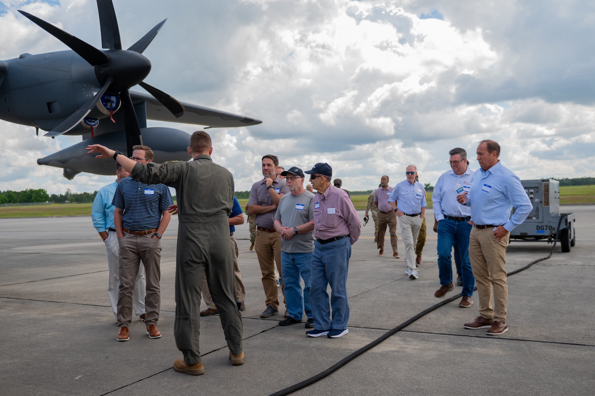 A group of people stand on a tarmac, listening to a pilot who is gesturing towards the plane.