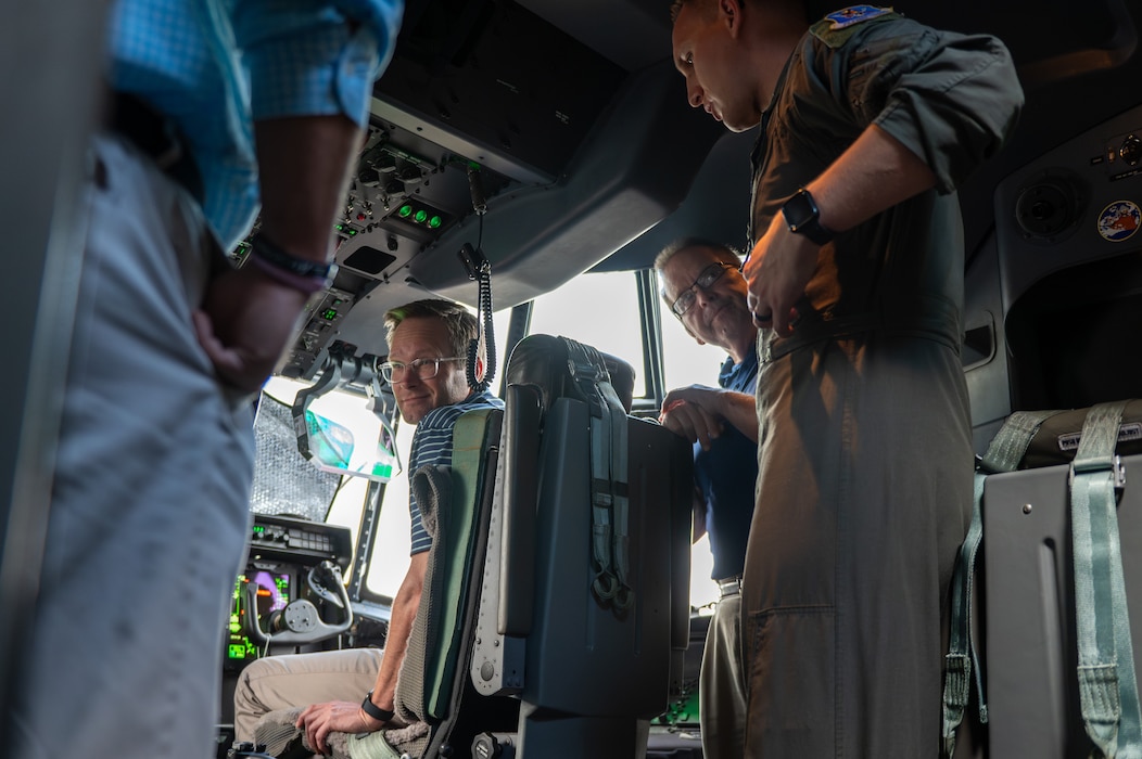 A man sits in a pilots chair, while another man stands behind him. Another man stands above them speaking to them.