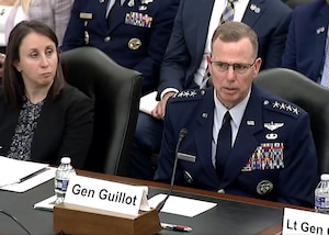 A man in a military uniform and a woman in business attire sit at a table speaking to a panel.