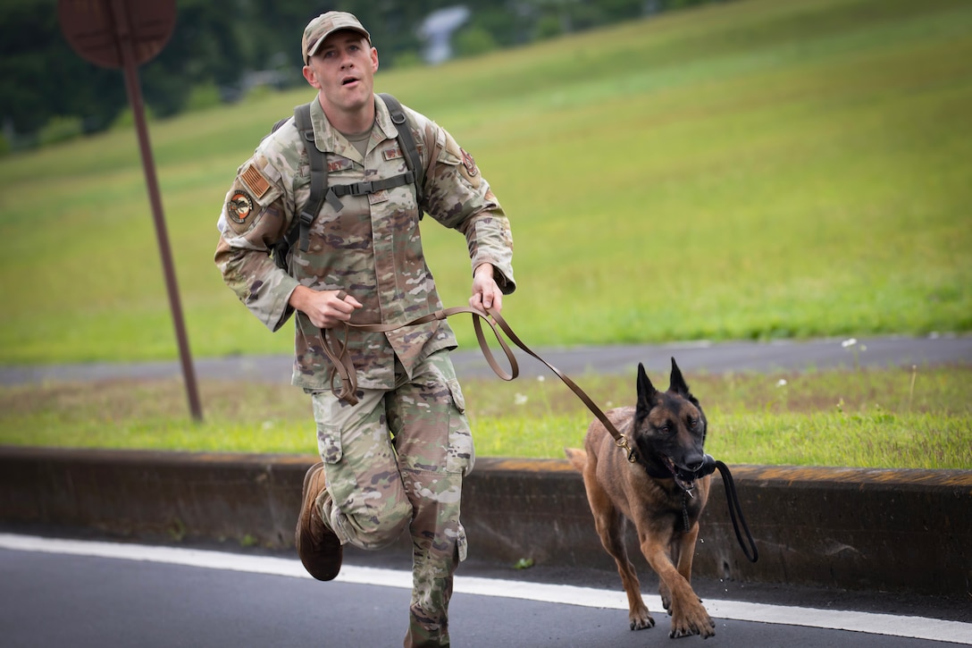 An airman and a military working dog run on pavement with a field in the background on a gloomy day.