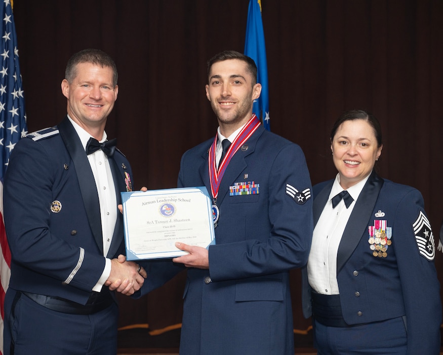 Two men stand to the left holding a certificate between them. A woman stands to the right. All of them are in uniform.