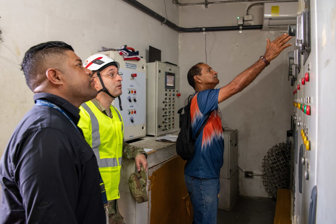 three men, one a Soldier, look at a panel of gauges and switches. The Soldier is wearing a hard hat and protective vest.