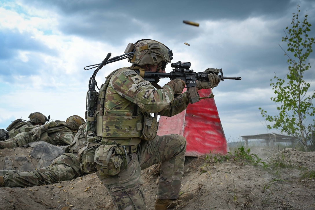 A soldier in tactical gear fires a weapon on a sandy hill alongside other soldiers on a cloudy day.