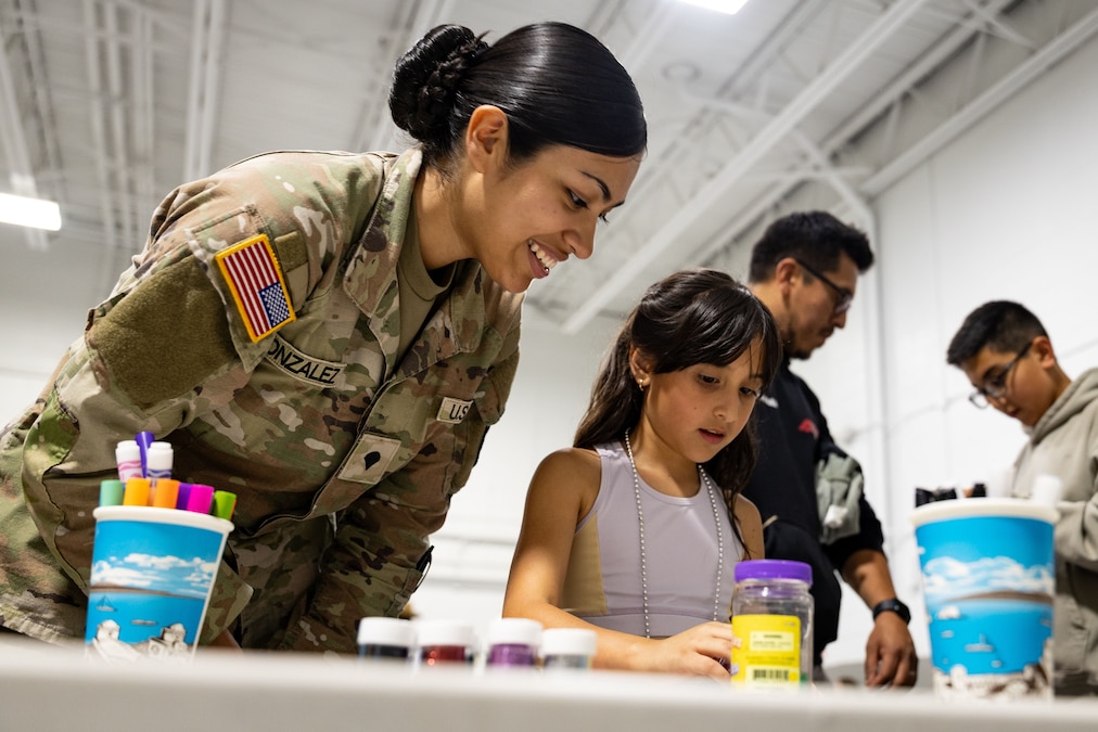 A soldier smiles as a child looks at a table with markers with two people in the background.