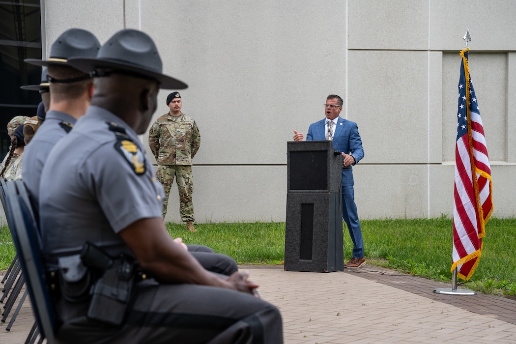 A male in a blue suit stands behind a podium and microphone beside a U.S. flag, state troopers sit in the foreground