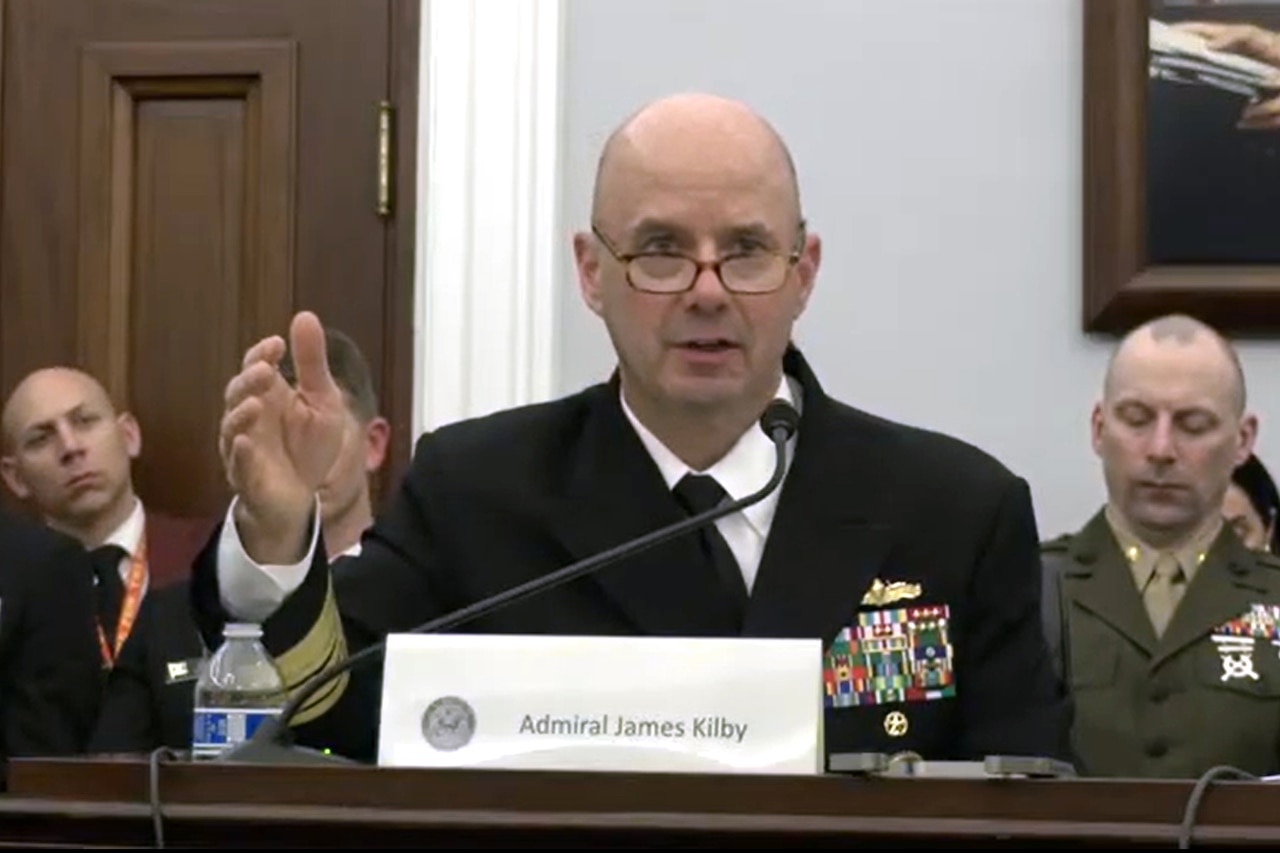 A man in a military dress uniform sits at a table and speaks into a microphone.