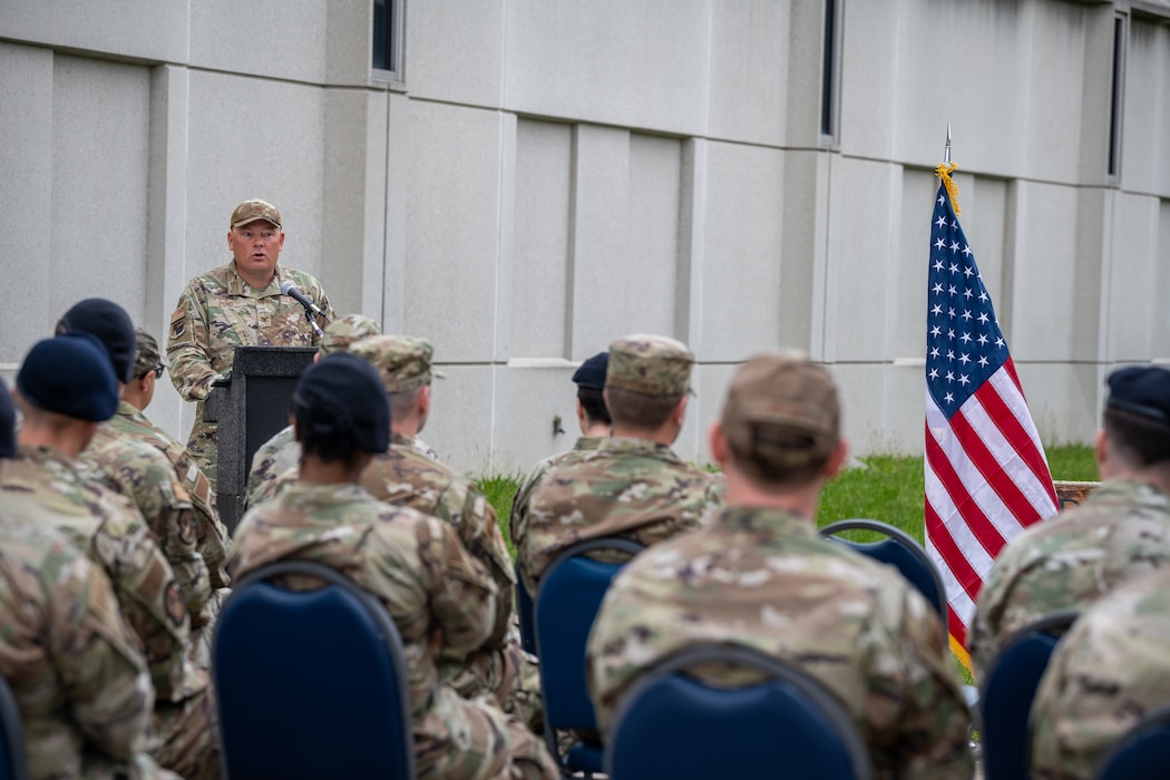 Col Pond stands behind a podium and microphone, seated attendees fill the foreground