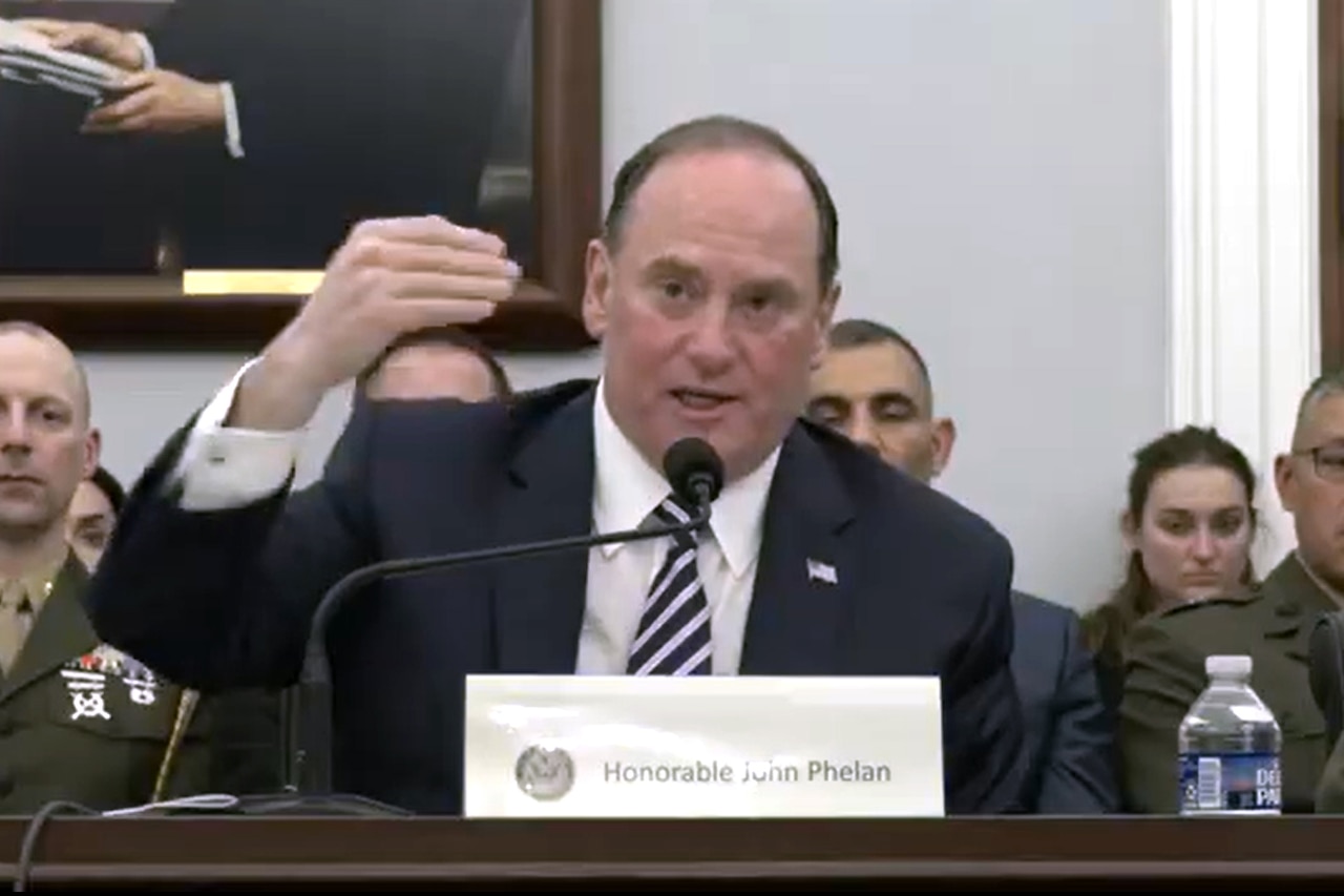 A man in a suit sits at a table and speaks into a microphone. Other people in military uniforms and business attire sit behind him.