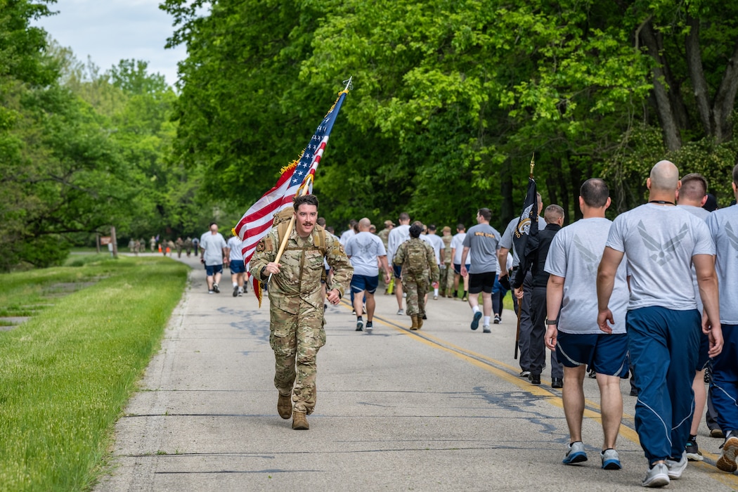 A male military member, wearing a camouflage uniform, carries a U.S. flag past a large group of marchers