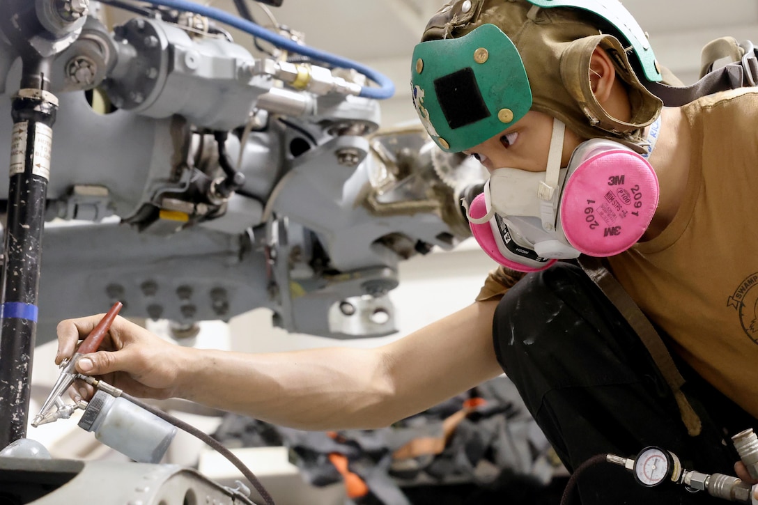 A sailor wearing a helmet and protective mouthpiece uses a tool to work on an aircraft.