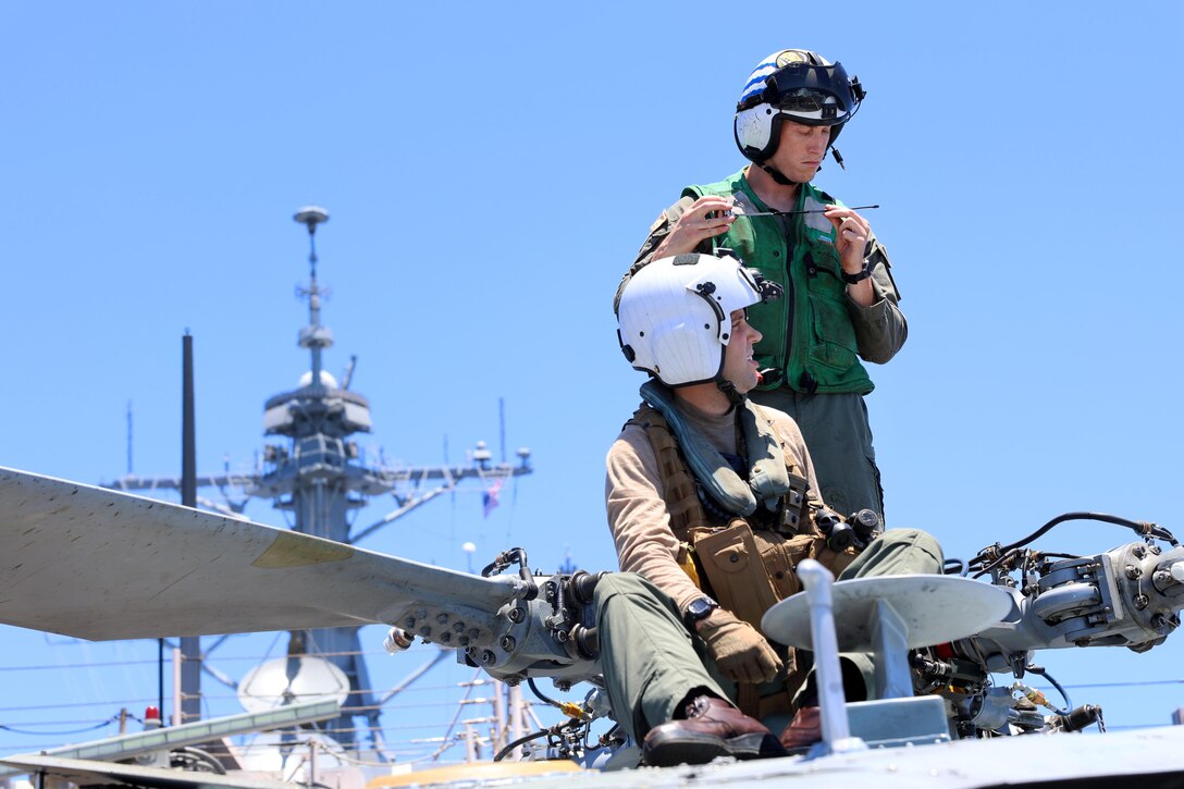 One sailor stands and another sits atop a parked helicopter on a ship, with blue sky in the background.