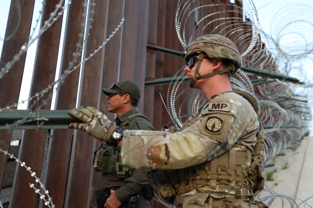A soldier and a border patrol again stand by a tall slated wall with barbed wire on it.