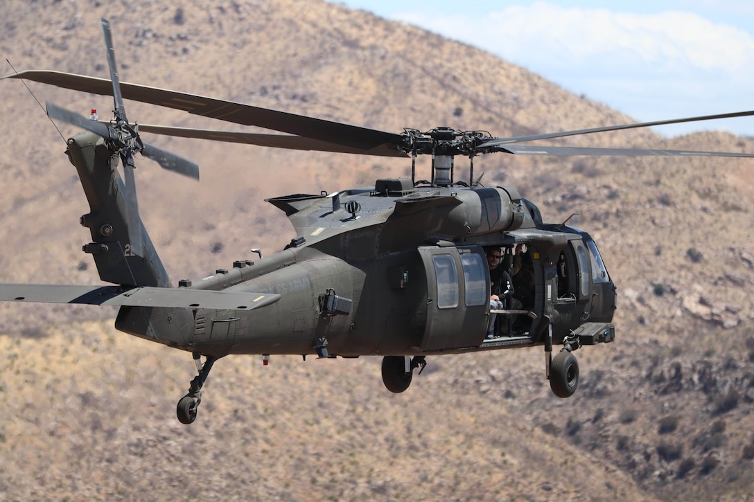 An Army helicopter flies over arid, hilly terrain.