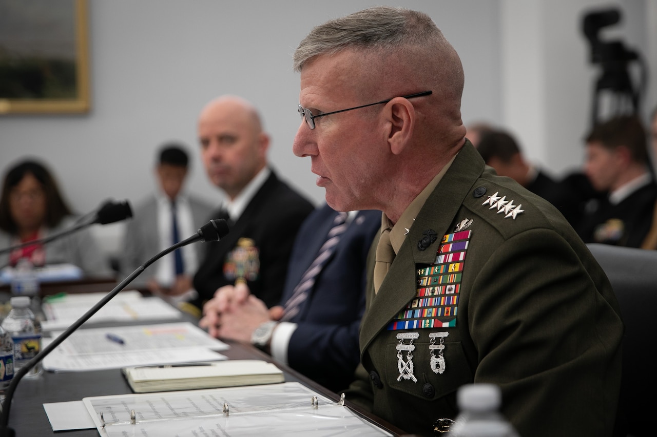 A man in a military dress uniform sits at a table and speaks into a microphone. Other people in military uniforms and business attire sit behind him. A woman in business attire is holding a camera.
