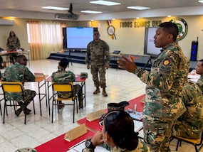A man in a military uniform stands and speaks to sitting military personnel.