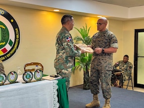 A man in a camouflage military uniform hand a plaque to another military person.