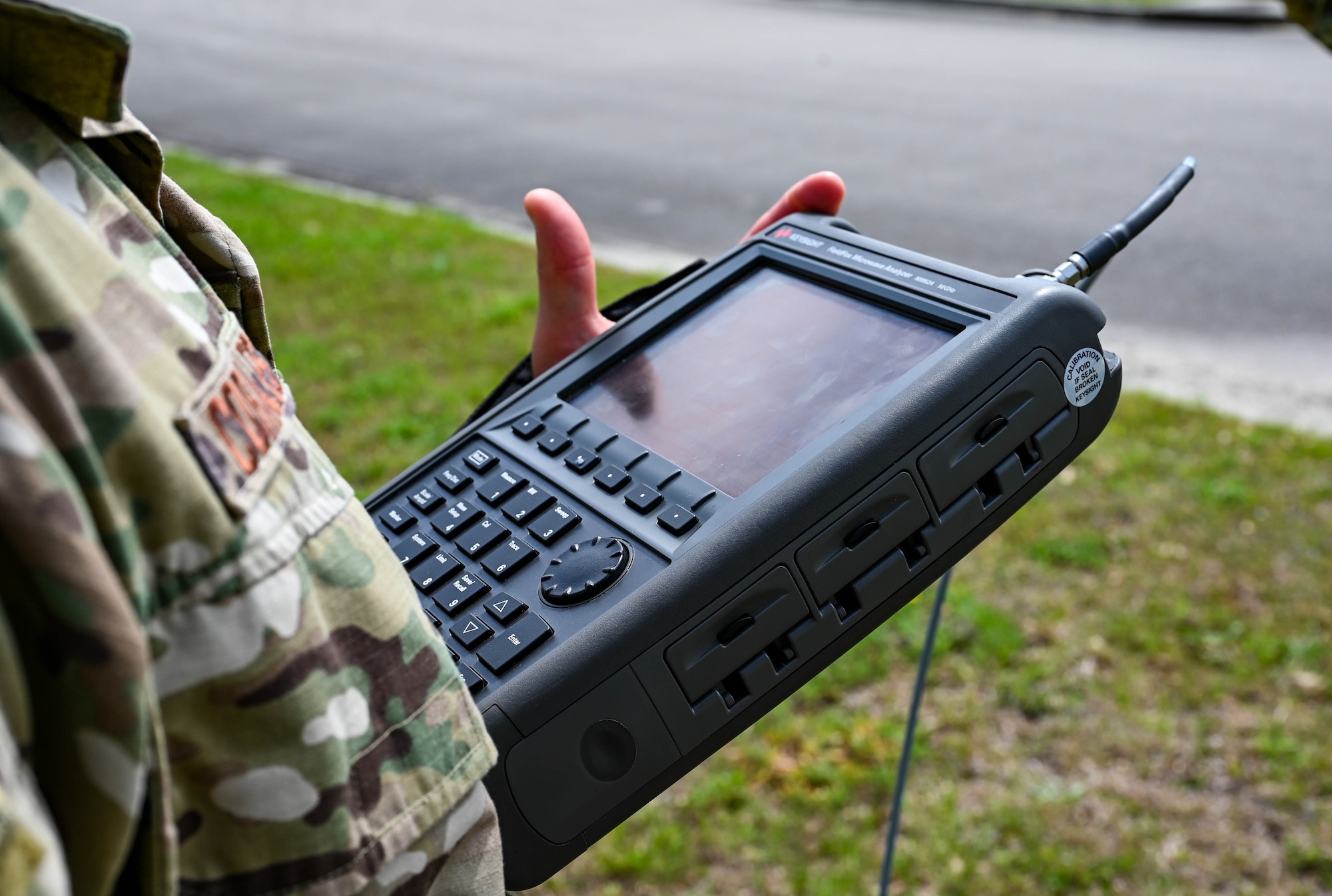 A close-up photo of an Airman holding a handheld radio frequency analyzer, looks like a large, thick calculator but bigger, with part of their camouflage uniform and name tape visible in the foreground.