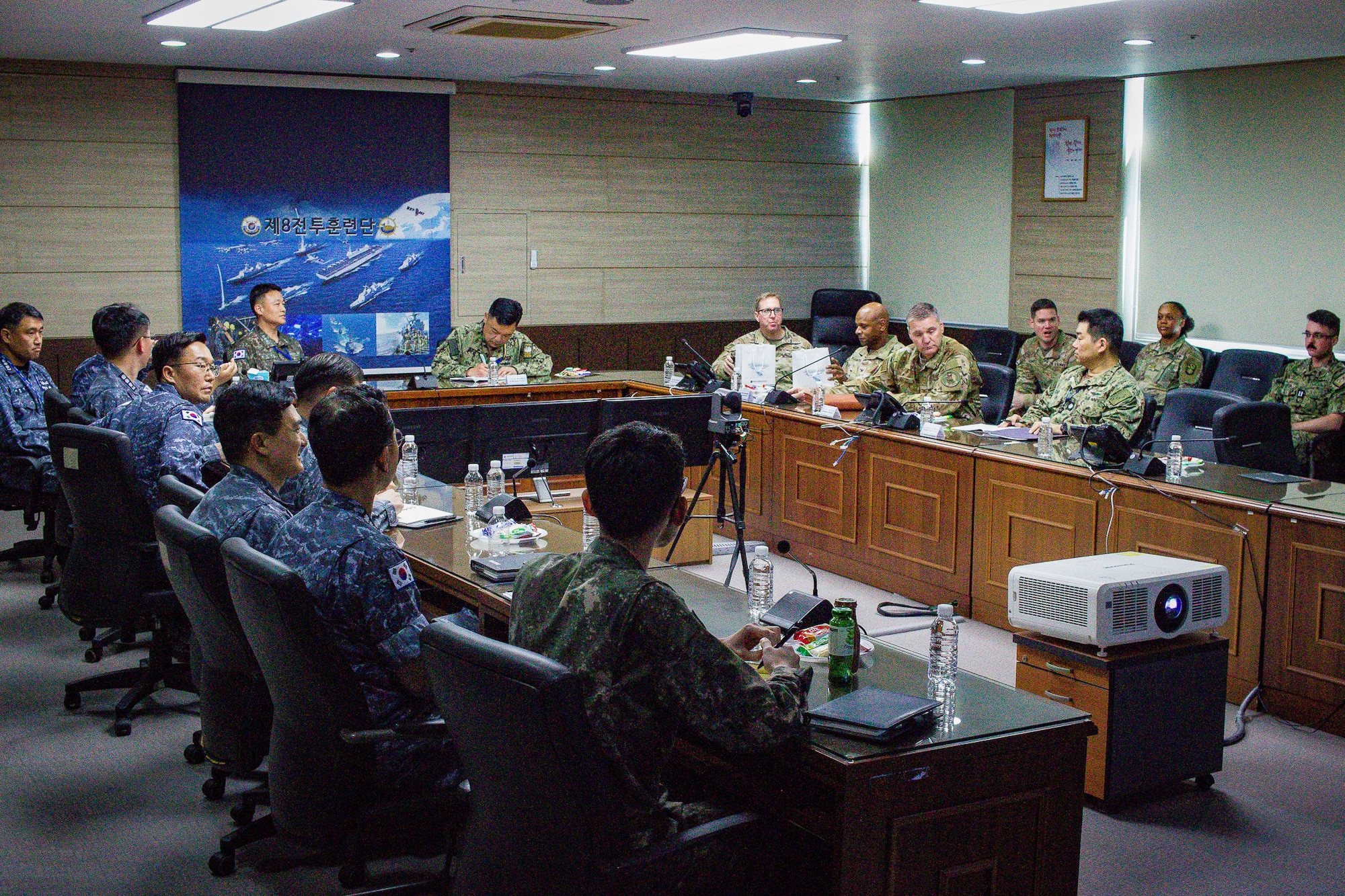 A U-shaped table has a dozen men in military uniforms staring at a screen