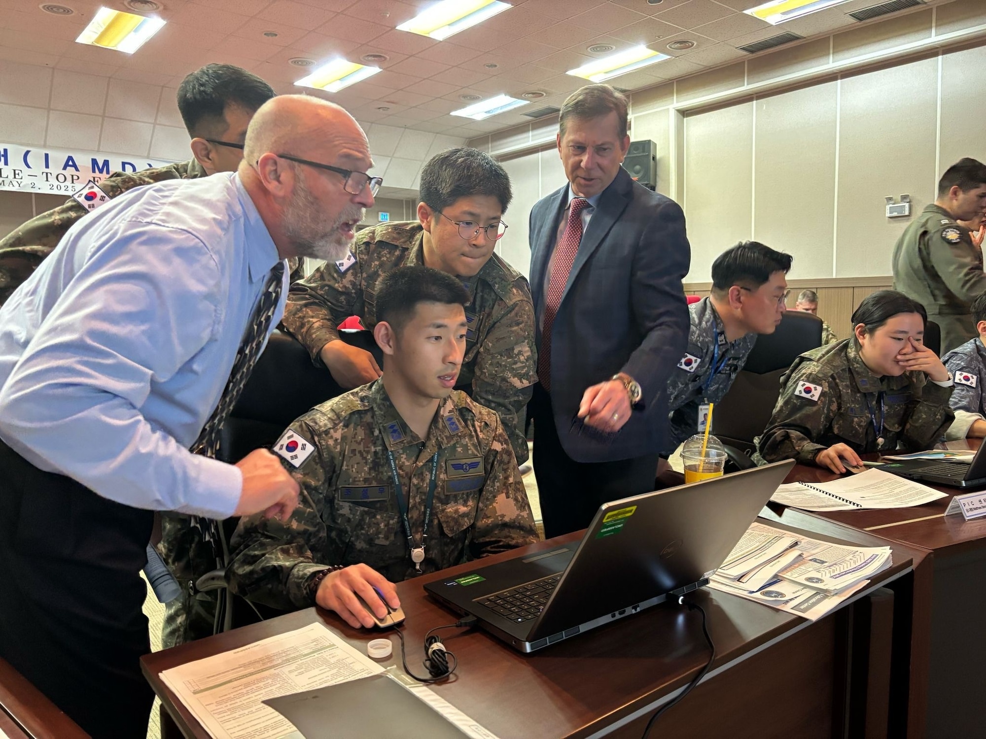Men stand around another man looking at a laptop on a long desk