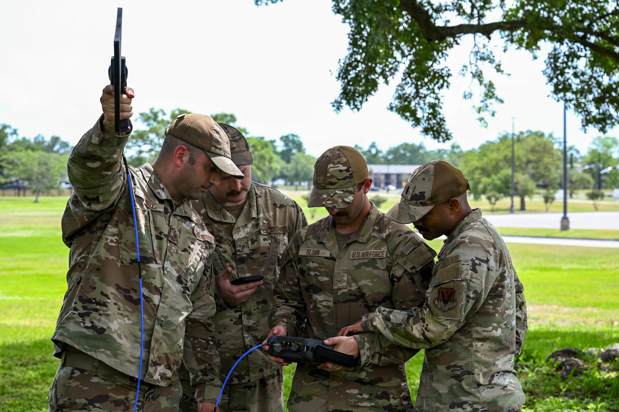 Four Airmen in military uniforms stand outside looking down at a radio frequency analyzer, that looks like a large, thick calculator but bigger and one holds a receiver in the air.