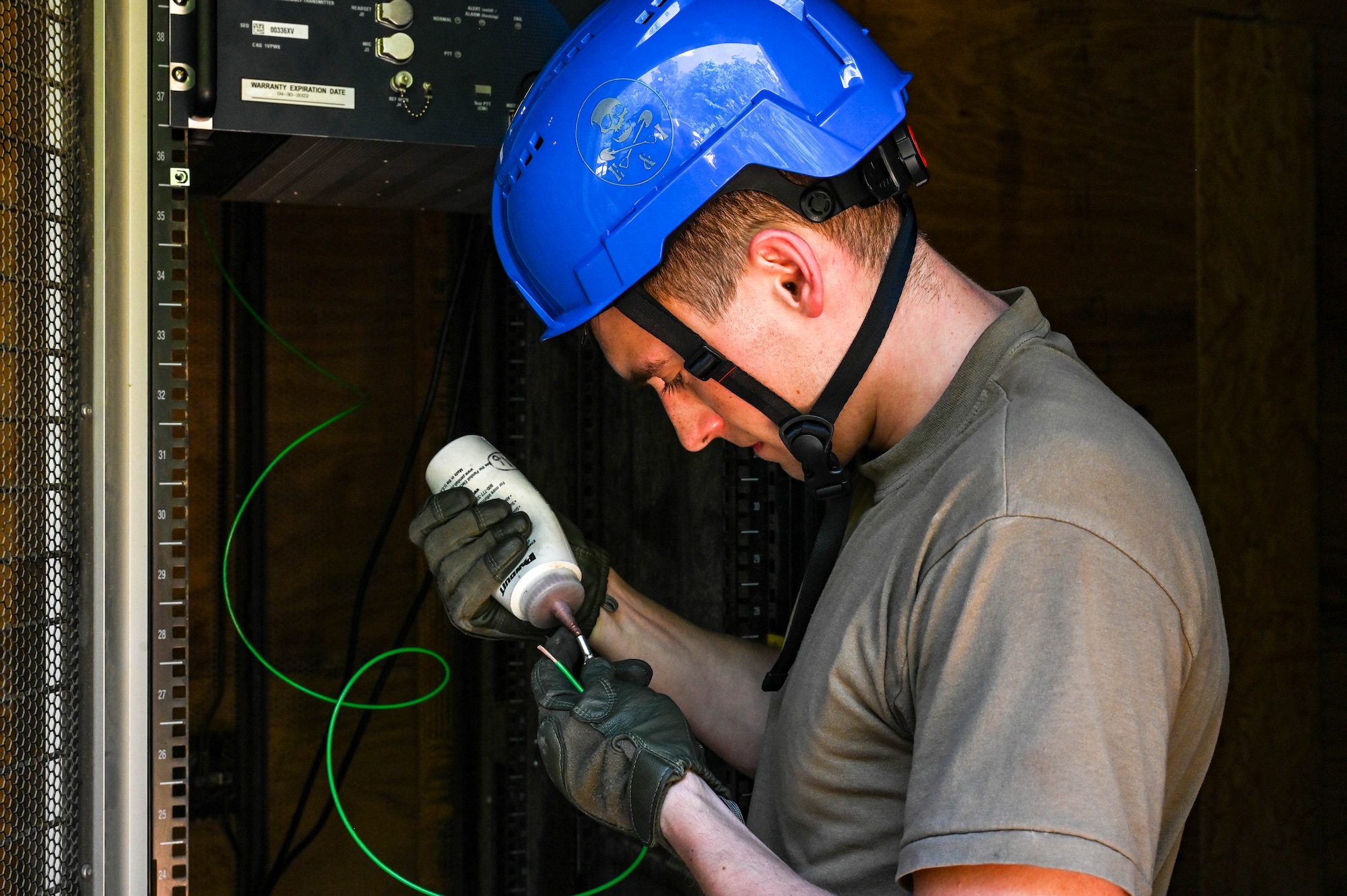 An Airman wearing a military uniform and a blue hard hat, applies anti-oxidation grease from a white squeeze bottle to a grounding lug.