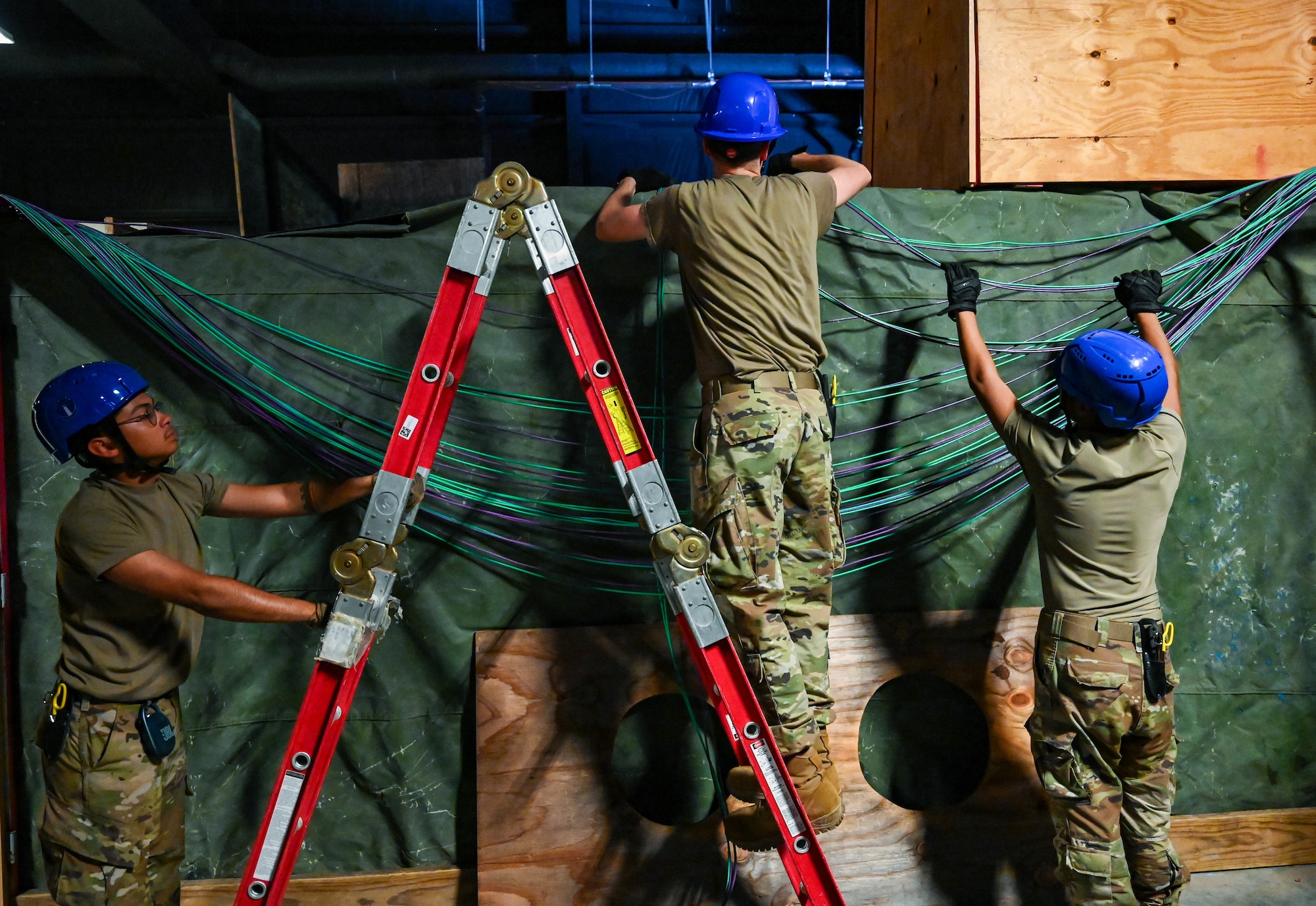 Three Airmen in military uniforms and blue safety helmets work together to install or organize a bundle of network cables along a green tarp-covered wall, while the one in the middle stands on a ladder