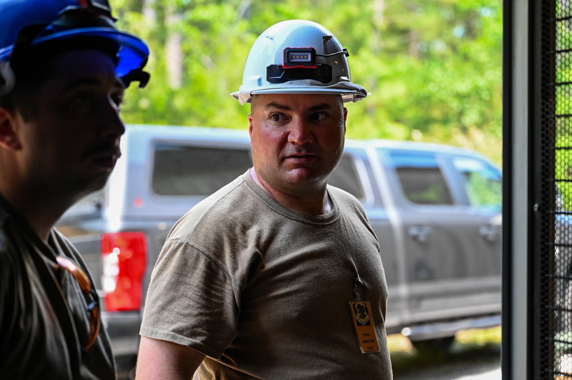 An Airman wearing a military uniform and white hard hat stands next to an Airman in a blue hardhat giving feedback during a field communications setup.
