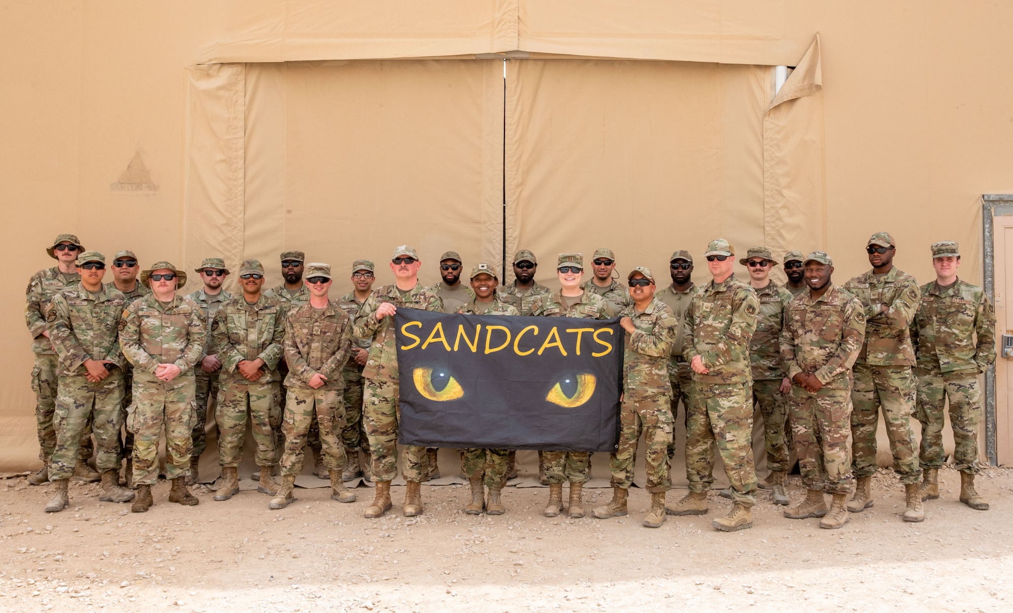 A group of Airmen pose for a photo with a black flag that has a pair of cat eyes on it and reads, Sand Cats.