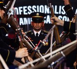 Army Soldiers dressed in dark ceremonial uniforms are performing a rifle drill routine while one Soldier walks up the middle of the formation.