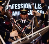 Army Soldiers dressed in dark ceremonial uniforms are performing a rifle drill routine while one Soldier walks up the middle of the formation.