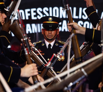Army Soldiers dressed in dark ceremonial uniforms are performing a rifle drill routine while one Soldier walks up the middle of the formation.