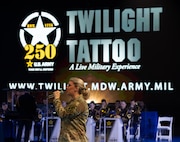 An Army soldier dressed in combat uniform is holding a microphone and singing while facing towards the left in front of a large display that reads Twilight Tattoo, a Live Military Experience
