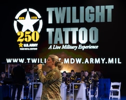 An Army soldier dressed in combat uniform is holding a microphone and singing while facing towards the left in front of a large display that reads Twilight Tattoo, a Live Military Experience