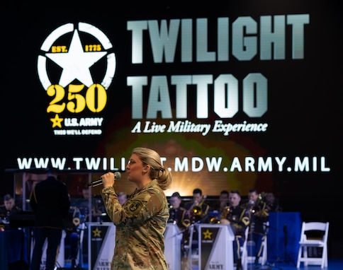 An Army soldier dressed in combat uniform is holding a microphone and singing while facing towards the left in front of a large display that reads Twilight Tattoo, a Live Military Experience