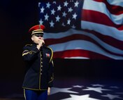 An Army Soldier dressed in dark ceremonial uniform with red hat is holding a microphone and singing while a large image of the US flag is projected in the background.