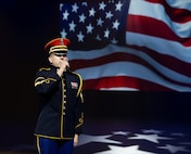 An Army Soldier dressed in dark ceremonial uniform with red hat is holding a microphone and singing while a large image of the US flag is projected in the background.