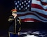 An Army Soldier dressed in dark ceremonial uniform with red hat is holding a microphone and singing while a large image of the US flag is projected in the background.