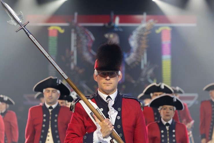 The U.S. Army Old Guard Fife and Drum Corps, wearing red Revolutionary War-era uniforms, is standing in formation during a Twilight Tattoo performance in Conmy Hall.