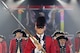 The U.S. Army Old Guard Fife and Drum Corps, wearing red Revolutionary War-era uniforms, is standing in formation during a Twilight Tattoo performance in Conmy Hall.
