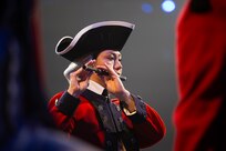 An Army Soldier dressed in Revolutionary War-era red uniform with black tri-cornered hat is playing a fife.