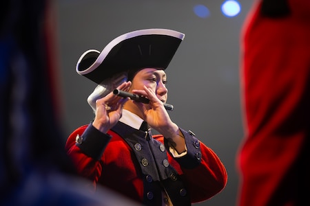 An Army Soldier dressed in Revolutionary War-era red uniform with black tri-cornered hat is playing a fife.